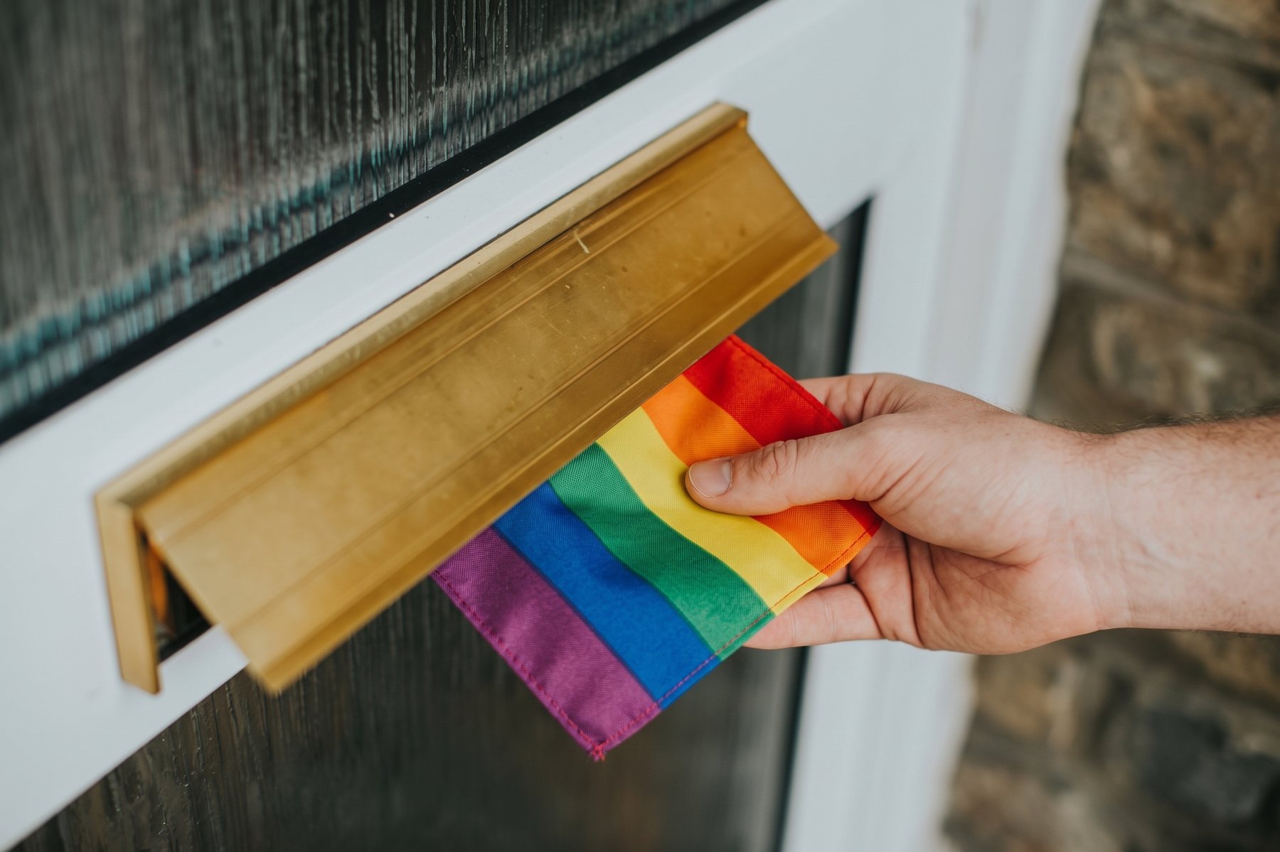Een hand duwt een regenboogvlag door de brievenbus van een voordeur