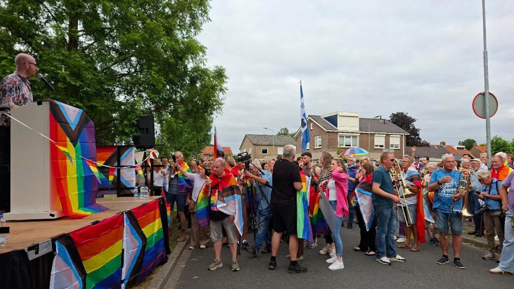Een spreker op een met regenboogvlaggen versierd podium richt zich tot een grote menigte