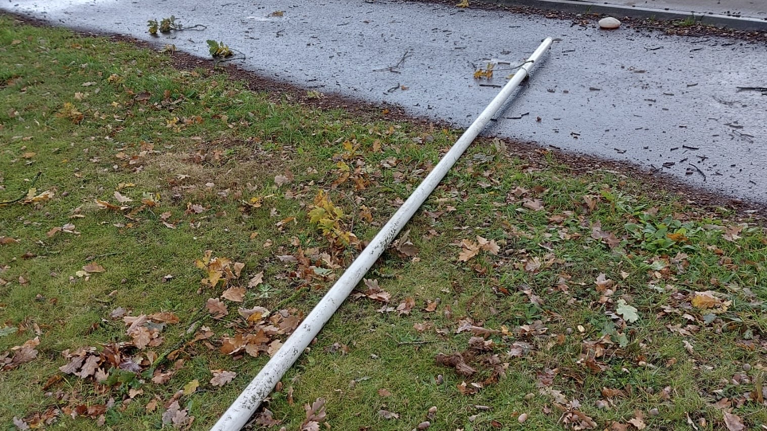 Een witte vlaggenmast ligt omvergetrokken in het gras langs een natte weg, tussen gevallen eikenblad